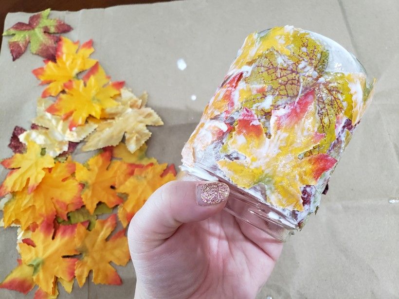 A hand holding a glass candle holder coated in fall leaves and mod podge 