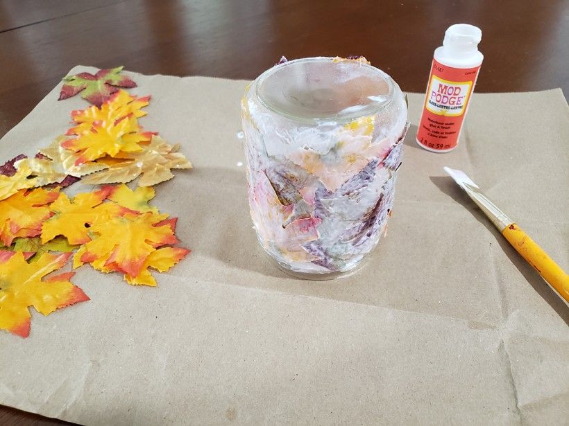 Mod podge covering a glass candle holder coated in fall leaves, with a paint brush, mod podge container, and extra leaves on a table