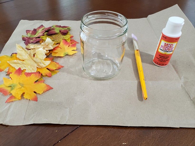 Project supplies, including a glass candle holder, paintbrush, mod podge, fall leaves, and a piece of brown paper on top of a table