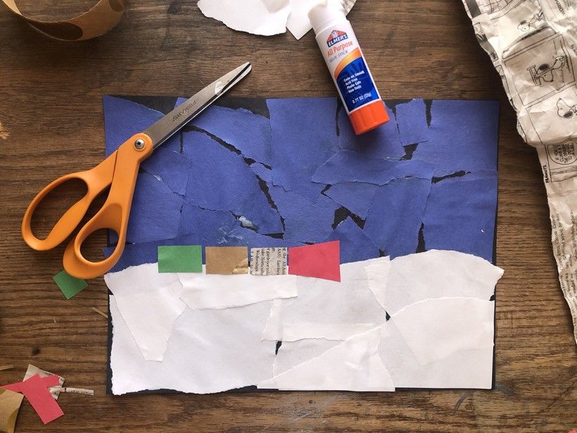 Colorful geometric shapes of construction paper glued over top of a background of ripped blue and white construction paper pieces. Scissors and glue stick are also in the frame.