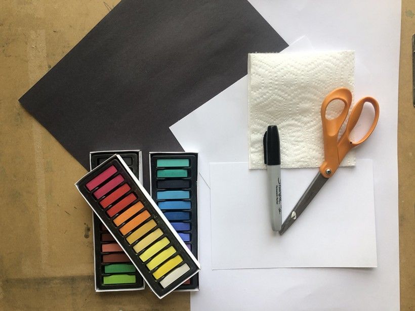 Photo of white and black pieces of paper, chalk pastels, a paper towel, scissors, and a sharpie