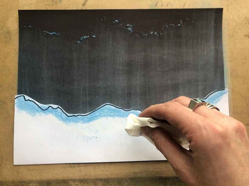 Black piece of paper with a piece of white paper on the bottom half and blue chalk pastels being colored on top