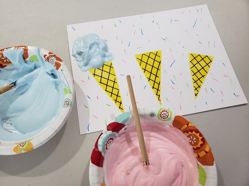 A white piece of paper with three yellow ice cream cones. One of the cones has a thick layer of blue puff paint on top to look like ice cream.