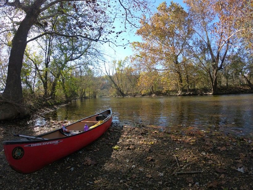 A red canoe on dry land next to a creek on a sunny autumn day