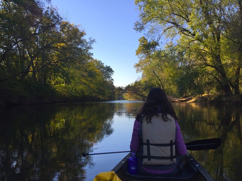 A woman wearing a life jacket and canoeing down the creek, lined with trees, on a sunny day
