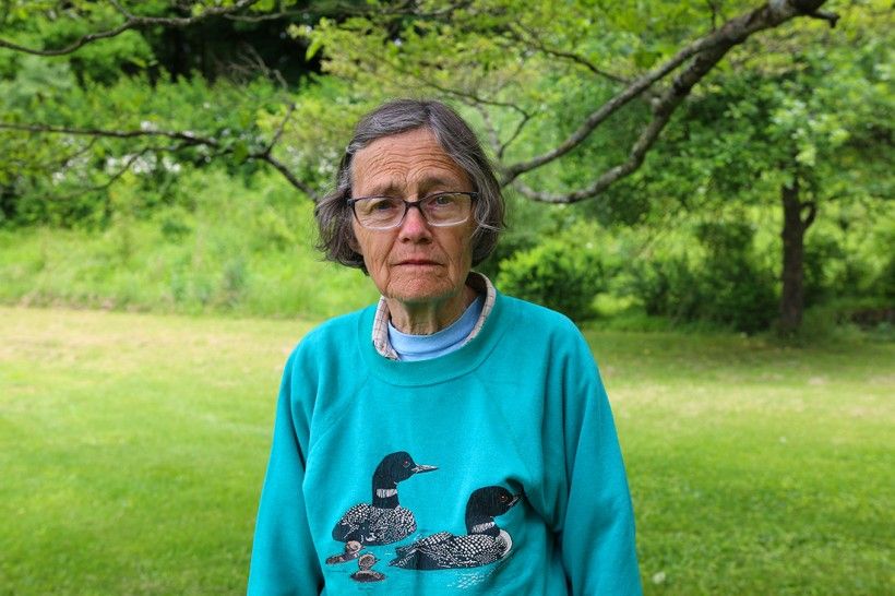 Photo of Janet Ebert, wearing a teal sweater with two ducks on the front. Photo is taken outside with green trees in the background.