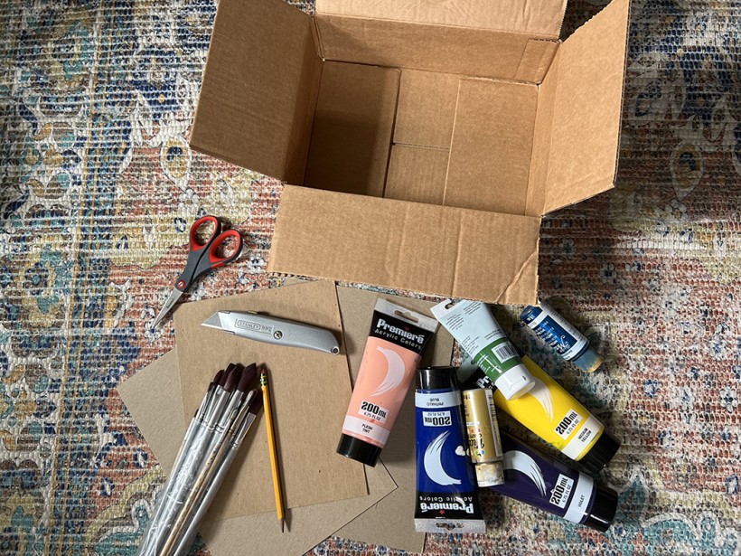 Shadowbox supplies: Empty cardboard box, scissors, various paints, paintbrushes, a few pieces of thin cardboard, and a razor cutter.