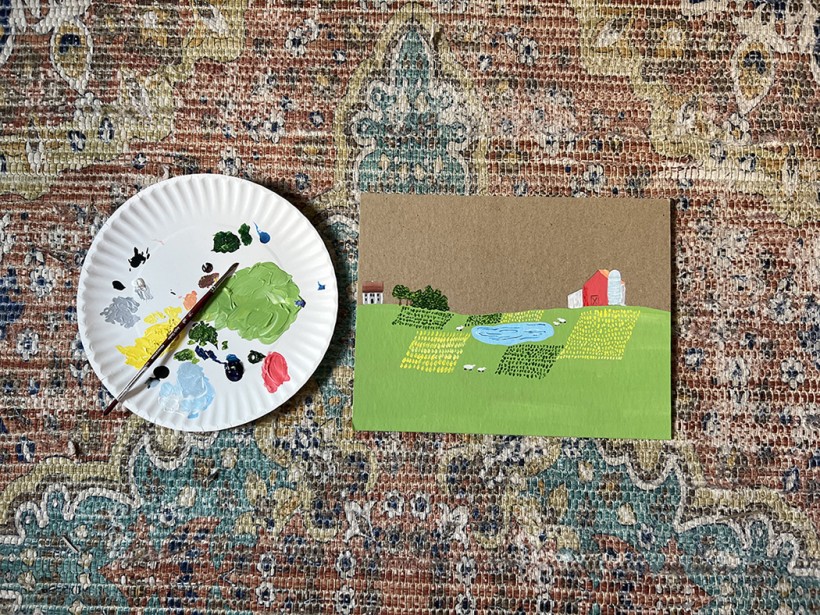 A white paper plate with an assortment of paint globs on top next to a thin piece of cardboard painted with a scene of a farm with a green, grassy hill, a pond, trees, and a red farmhouse on top of the hill.