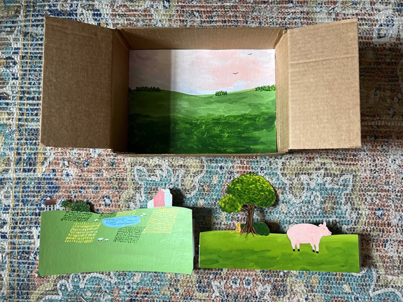 An open cardboard box with a painted pastoral landscape scene at the bottom of the box. Two cutout landscape scenes are cutout and ready to be added to the open cardboard box.