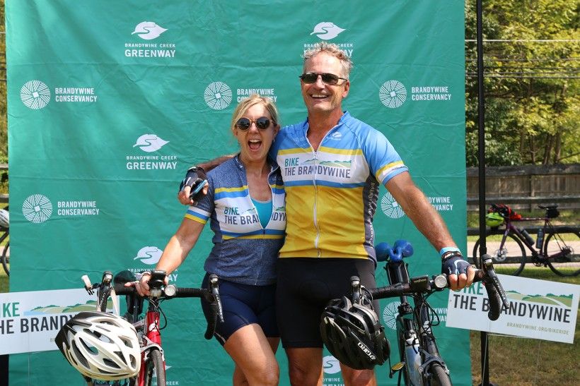 Photo of two Bike the Brandywine riders in front of a green step-and-repeat backdrop with the Brandywine Conservancy and Brandywine Creek Greenway logos