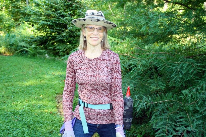 Photo of Margaret Moore, wearing a hat, long-sleeved shirt, garden gloves, and a utility belt while standing in her woodlands.