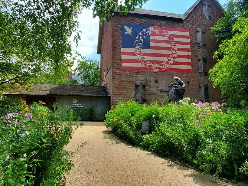 James Prosek (b. 1975), <em>Invisible Boundaries,</em> 2022, digital print on polyester scrim vinyl, 16' x 27'. Courtesy of the artist and Waqas Wajahat, NY. Photograph courtesy of the Brandywine River Museum of Art. Front entrance of the Museum with a large artwork installed on the mill building.