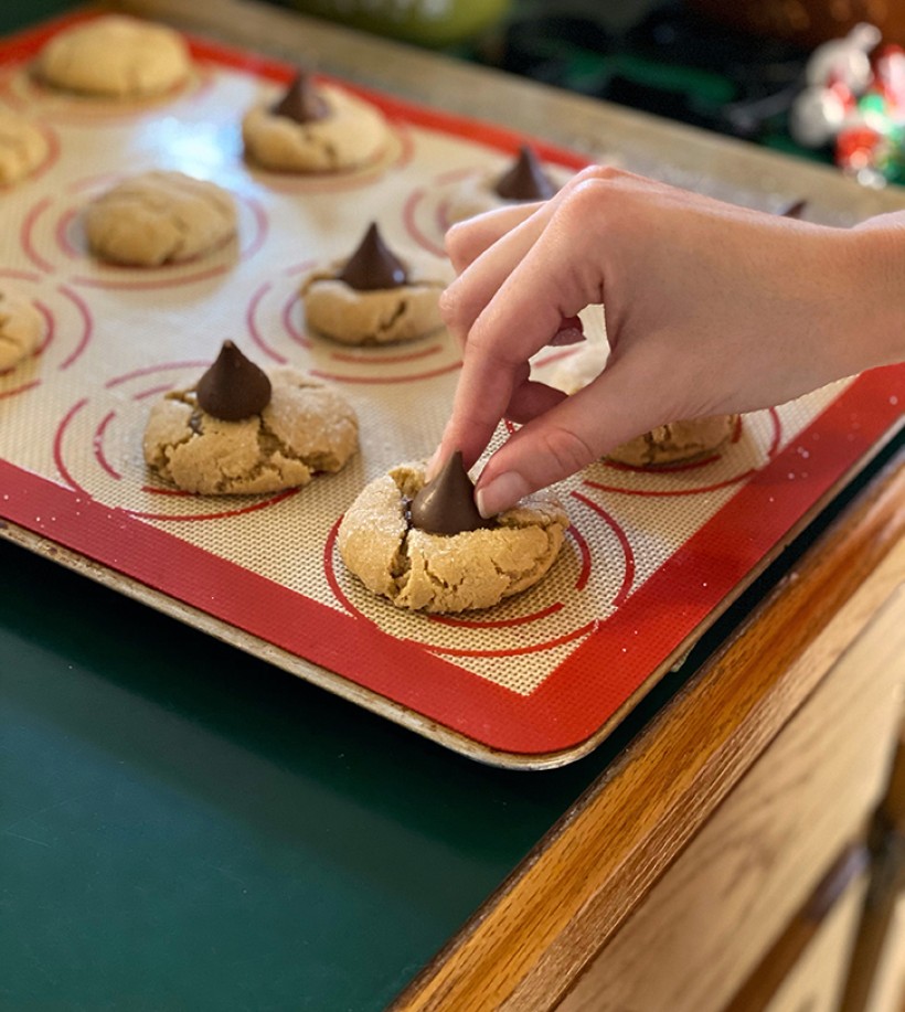 Peanut butter blossom cookies on a cookie sheet with a hand placing a Hershey kiss chocolate on top of a baked cookie