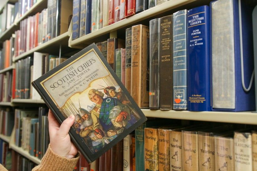 A hand holding a book next to a large shelf of books.