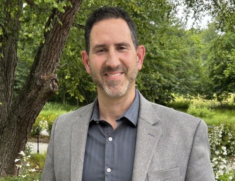 a man in a grey suit standing in front of lush green trees and flowers