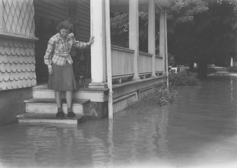  A historical black and white photograph, taken in 1942, showing a woman identified as Anna Seal Schlosser standing on the steps of her home's porch immediately after a flood in Chadds Ford. She is wearing a plaid shirt and a dark skirt, looking down at the steps. The porch has white columns and a railing. Floodwater covers the entire yard, reaching the second step and soaking the wooden lattice foundation of the porch.