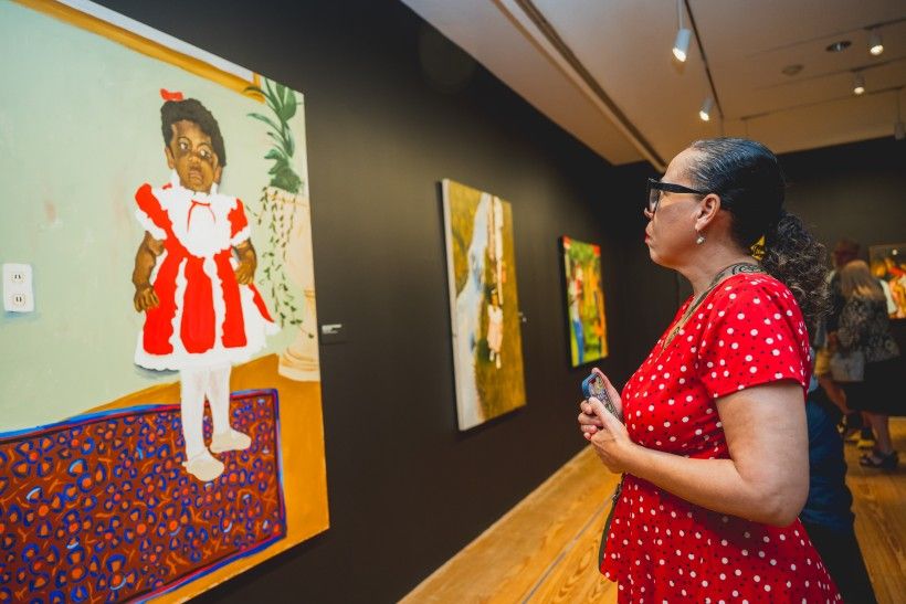 On the right, a woman with glasses and her dark hair pulled back stands in profile, looking intently at a painting on the wall to her left. She is wearing a bright red dress with white polka dots and holding a smartphone. The closest painting is a portrait of a young Black girl wearing a red and white ruffled dress, standing on a patterned blue and red rug.