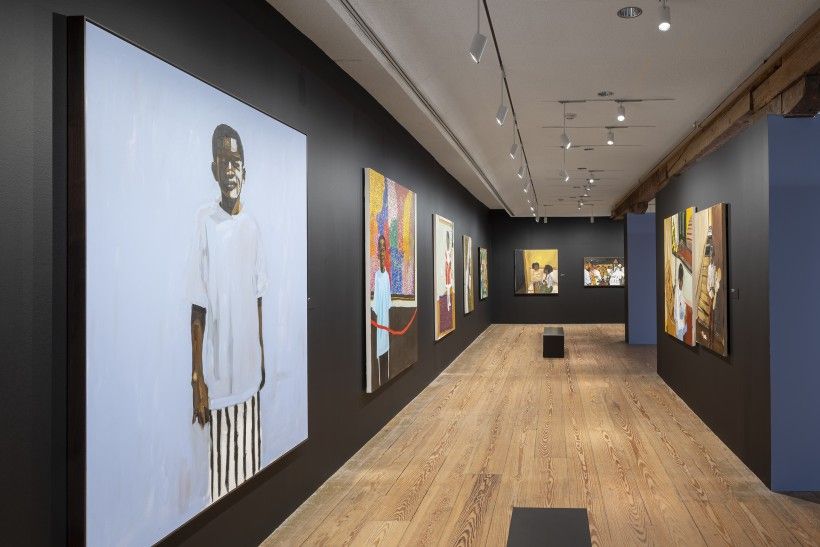  A photograph capturing a long, narrow gallery hallway with wide wooden floorboards and dark gray walls. Large, brightly colored figurative paintings are hung along both walls. 