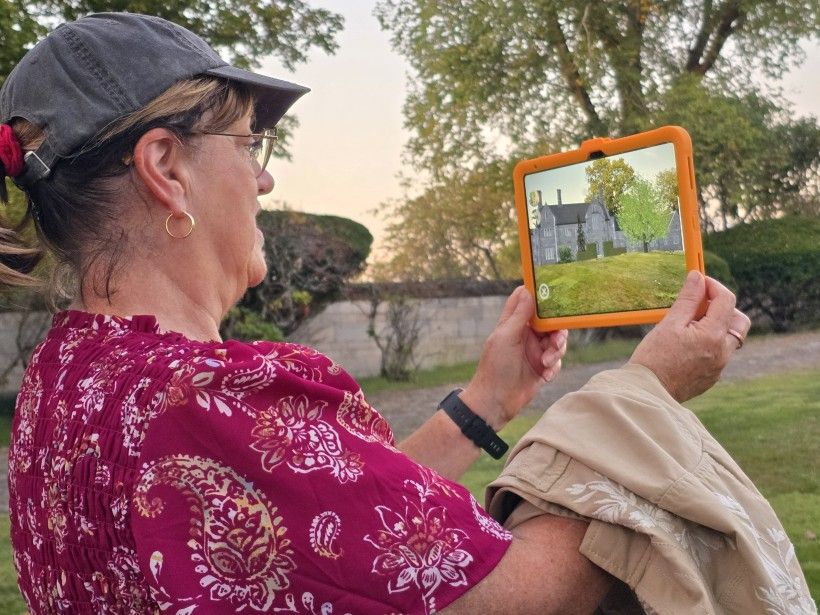 a photograph of a woman holding a digital tablet to view an augmented reality rendering of a mansion that once stood on the site