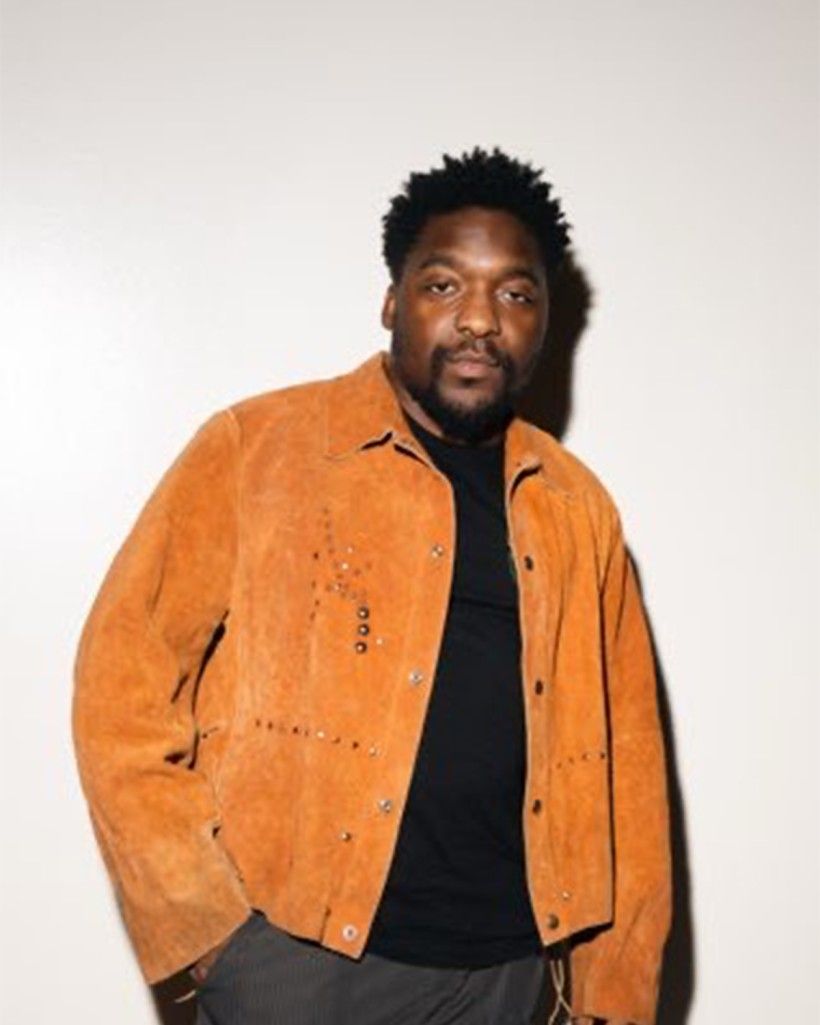 This image shows a man posing for a photo, standing against a plain white wall. He is facing the camera directly with a calm, confident expression. He is wearing a bright orange colored jacket, with a black shirt and gray trousers.