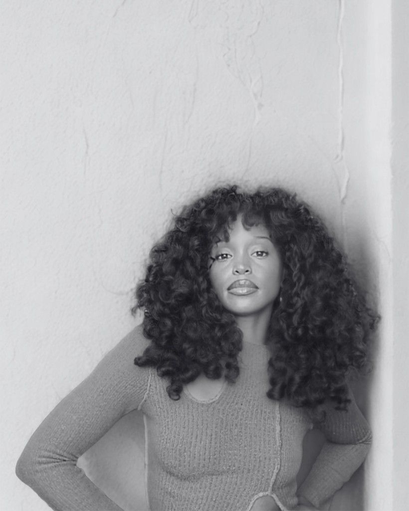 This is a black-and-white portrait of a woman standing against a plain, textured wall. She is facing the camera directly with a calm, confident expression. She has long, full, curly hair that frames her face and falls around her shoulders.