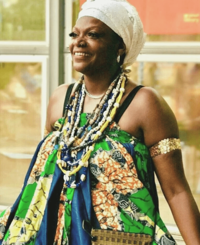 A smiling woman stands indoors in front of large windows that let in soft daylight. She appears confident and joyful. She is wearing a colorful, patterned dress made of African-style fabric in green, blue, yellow, and orange tones. The dress is bright and flows loosely.