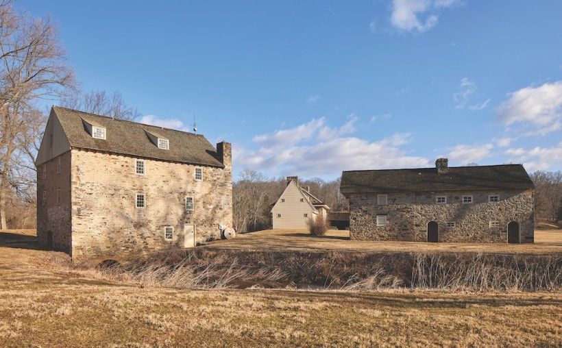 A group of old stone buildings sits in an open grassy field under a bright blue sky with a few scattered clouds. The largest structure has thick stone walls and a steep roof, while a smaller barn and another house stand nearby, giving the scene a quiet, historic countryside feel.