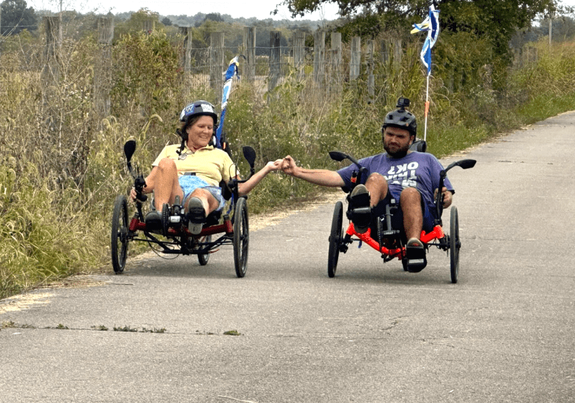 Two people are riding low, three-wheeled recumbent bicycles side by side on a quiet paved country road bordered by tall grass and wooden fence posts. They are reaching out to hold hands while riding, wearing helmets and smiling.