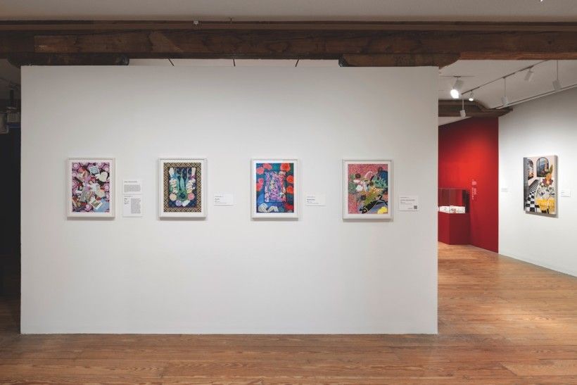 A clean, well-lit art gallery shows several colorful framed paintings arranged evenly on a large white wall. Wooden floors and beams, along with a red accent room in the background, give the space a modern yet warm feel.
