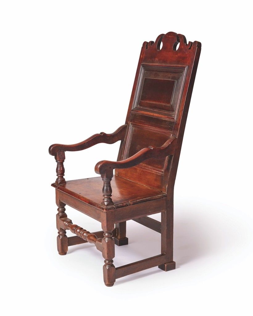 A tall, dark wooden armchair is shown against a plain white background, with a high decorative back and curved armrests. Its sturdy frame has carved legs, turned supports, and a horizontal footrest connecting the front legs.
