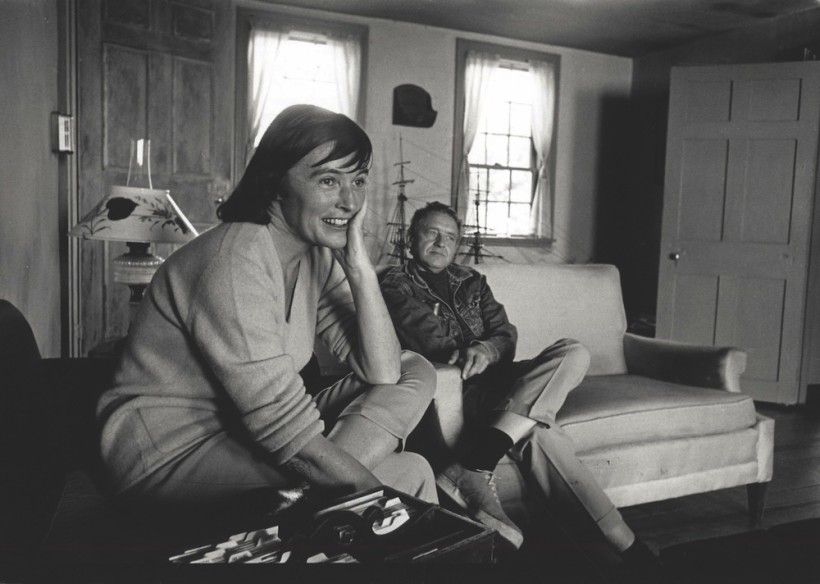 A black-and-white photograph shows two people relaxing in a cozy living room, with one person seated in the foreground smiling and resting their head on their hand. The other person sits casually on a couch behind them, surrounded by a lamp, windows, a model ship, and simple wooden furnishings.