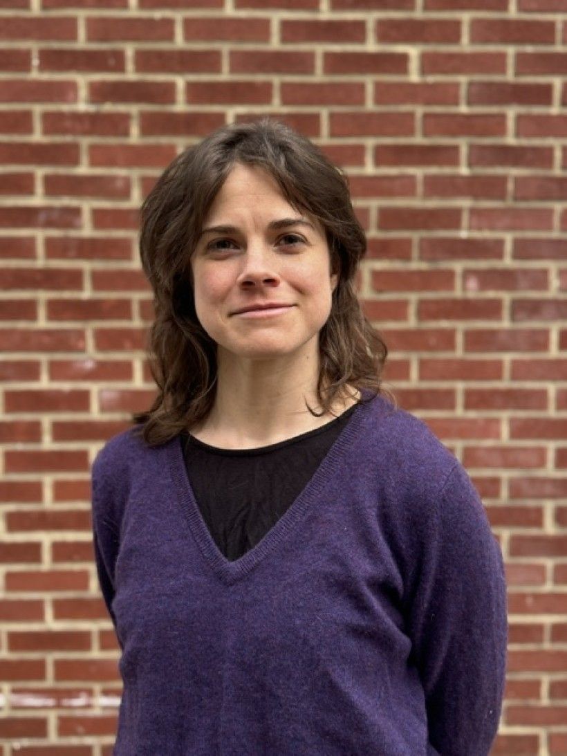 Woman standing in front of a brick wall, smiling at the camera, wearing a purple sweater