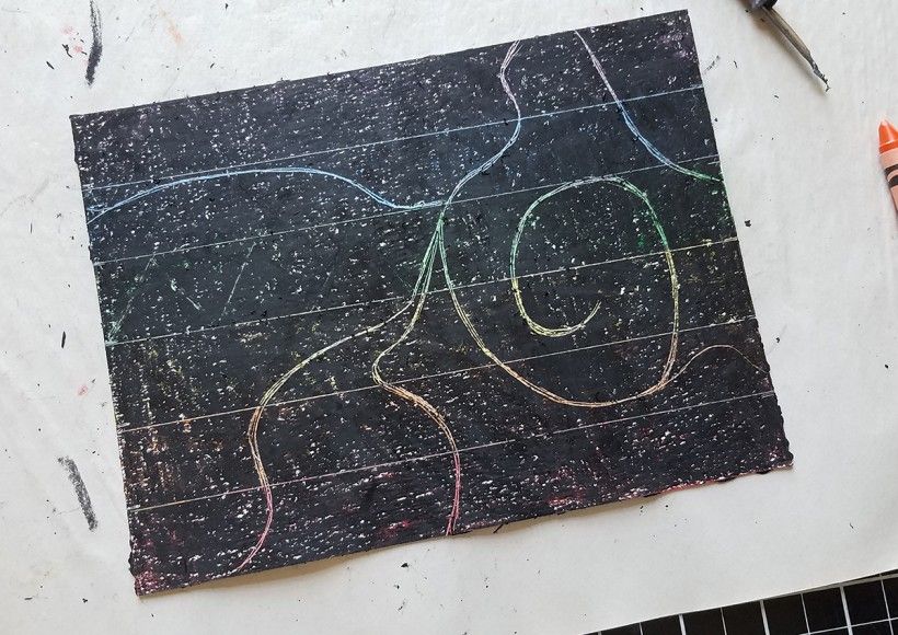 A piece of paper colored over with black crayon, with a colorful design scratched through the middle of the paper
