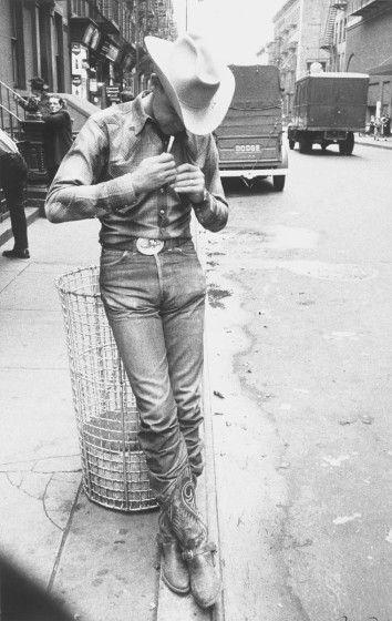 A black and white shot of a cowboy standing against a trash can and lighting a cigarette on a city street.