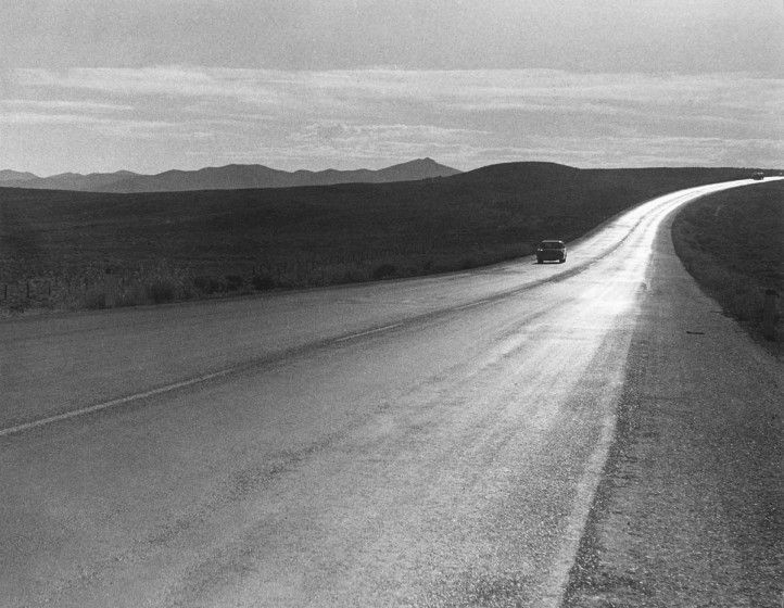 A black and white shot of an open road from the perspective of the side of the road with mountains in the distance.