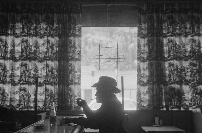 A black and white image of a silhouette of a man in a cowboy hat in front of a window at a diner.